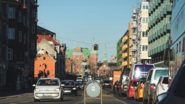 En travl bygade med biler, der holder for rødt ved et trafiklys, farverige bygninger på begge sider og en klar, blå himmel over dem.