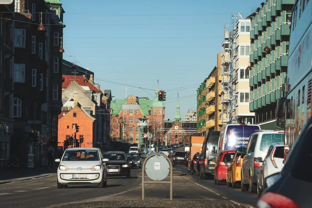 En travl bygade med biler, der holder for rødt ved et trafiklys, farverige bygninger på begge sider og en klar, blå himmel over dem.