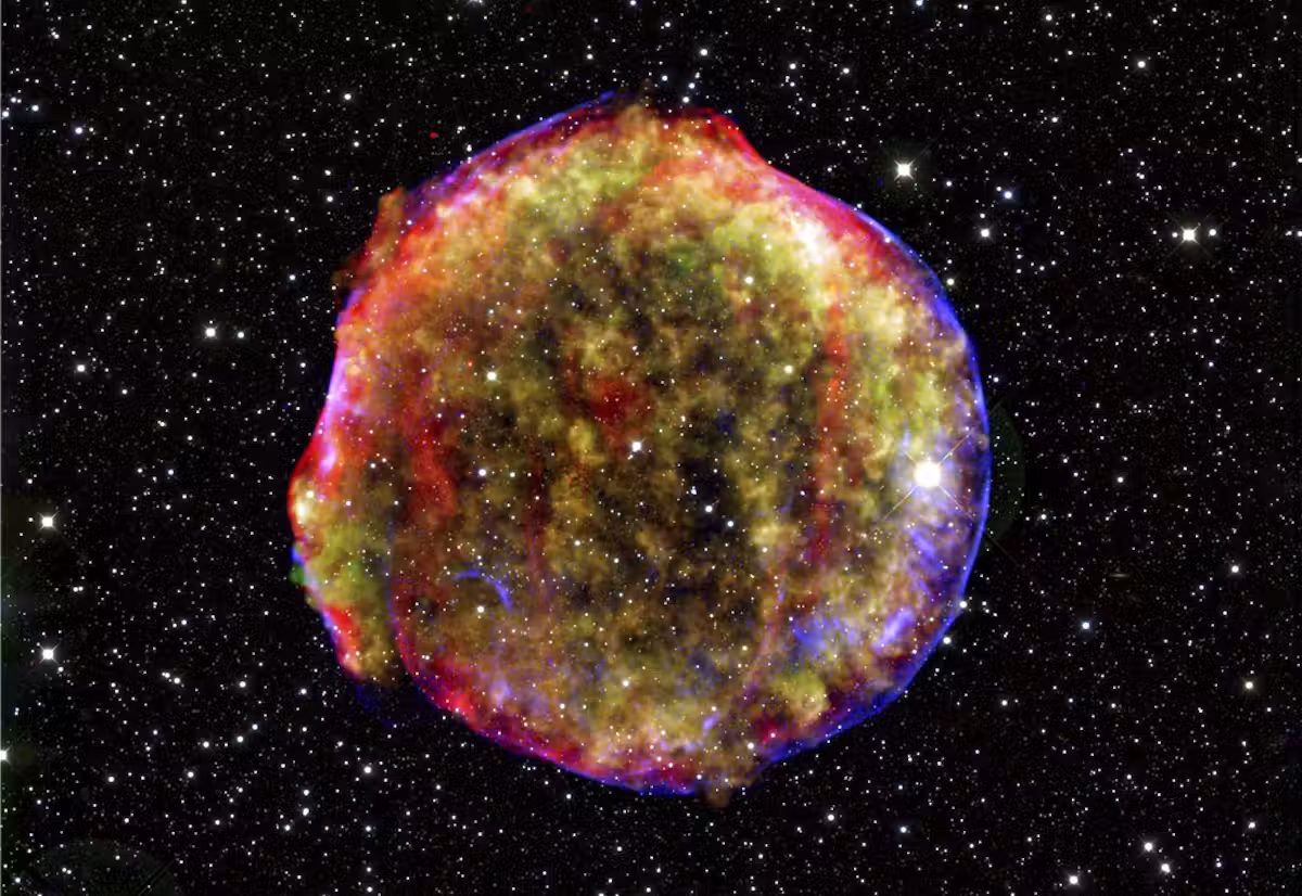 En farverig, lysende supernovarest, der ligner en tåge, med røde, gule, grønne og blå nuancer udbreder sig på baggrund af utallige små hvide stjerner i rummets mørke.