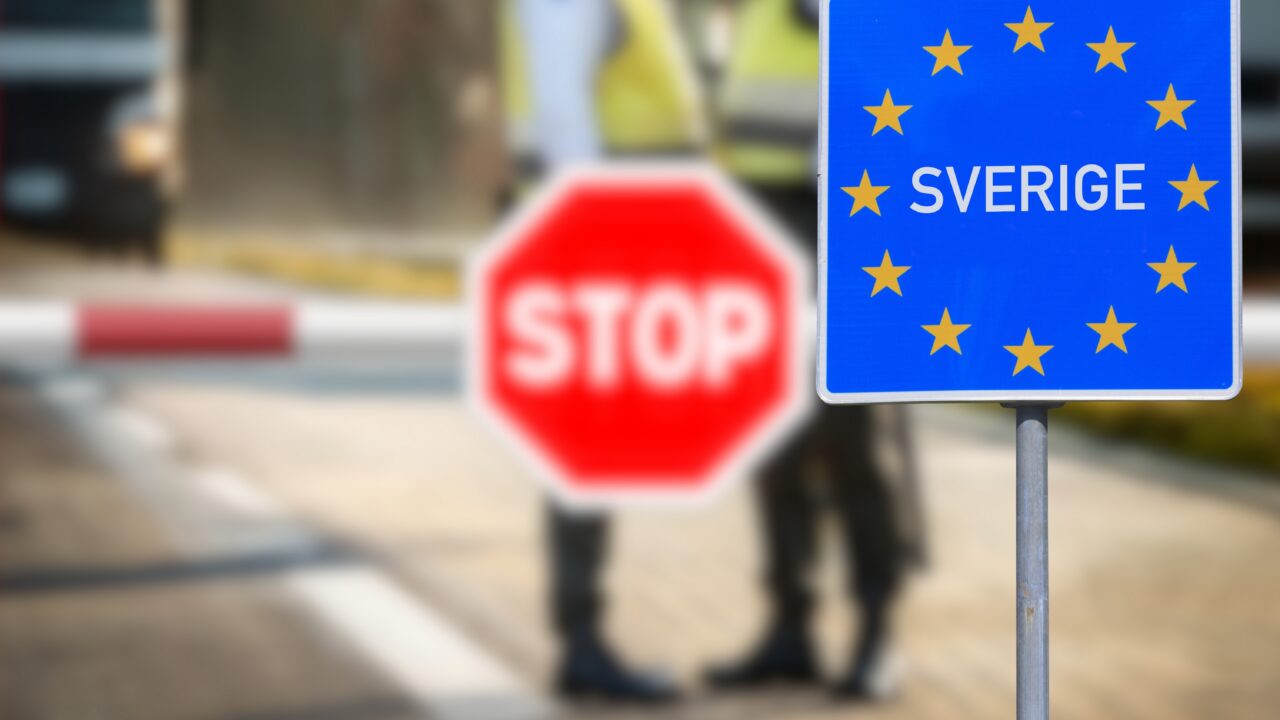 Et blåt skilt med gule EU-stjerner og teksten »SVERIGE« markerer den svenske grænse. Bag skiltet ses et rødt STOP-skilt og to personer i uniform, der er lidt slørede.