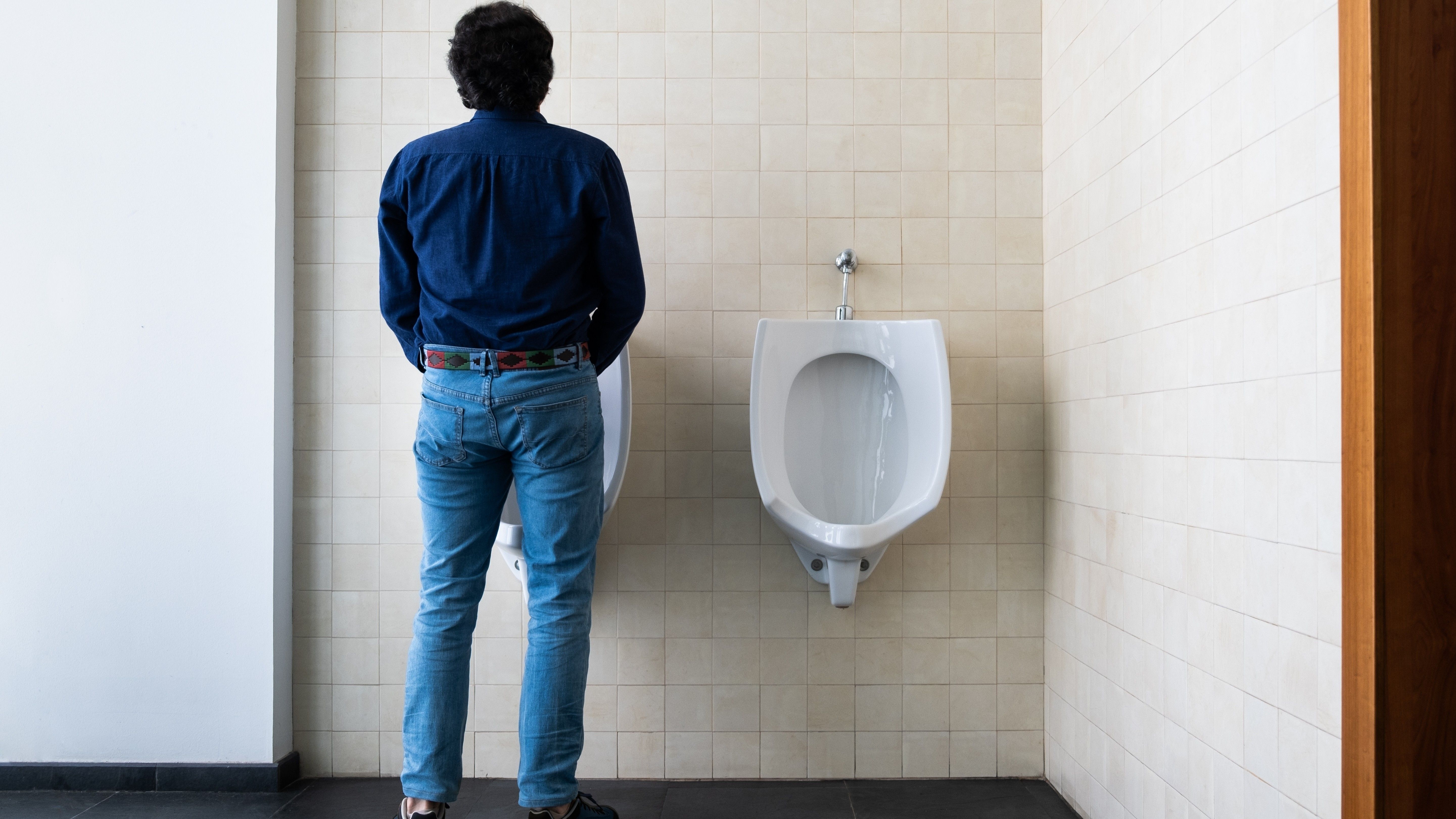 En person iført en mørk skjorte og blå jeans står vendt mod et hvidt urinal på et offentligt toilet med beige flisebelagte vægge.