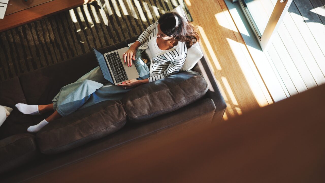 En kvinde sidder på en brun sofa og arbejder på en bærbar computer. Hun har en stribet trøje, en hvid bluse, blå jeans og hvide sokker på. Sollyset strømmer ind gennem de store vinduer og kaster skygger på gulvet og tæppet.