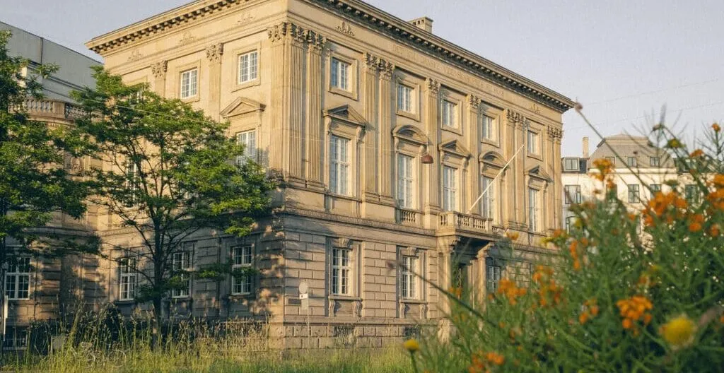 En historisk, beige stenbygning med høje vinduer og søjler står bag træer og vilde blomster på en solskinsdag.