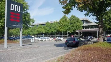 Et stort DTU-skilt og -logo tæt på en parkeringsplads fyldt med biler, træer og en moderne bygning i baggrunden på Danmarks Tekniske Universitets campus.