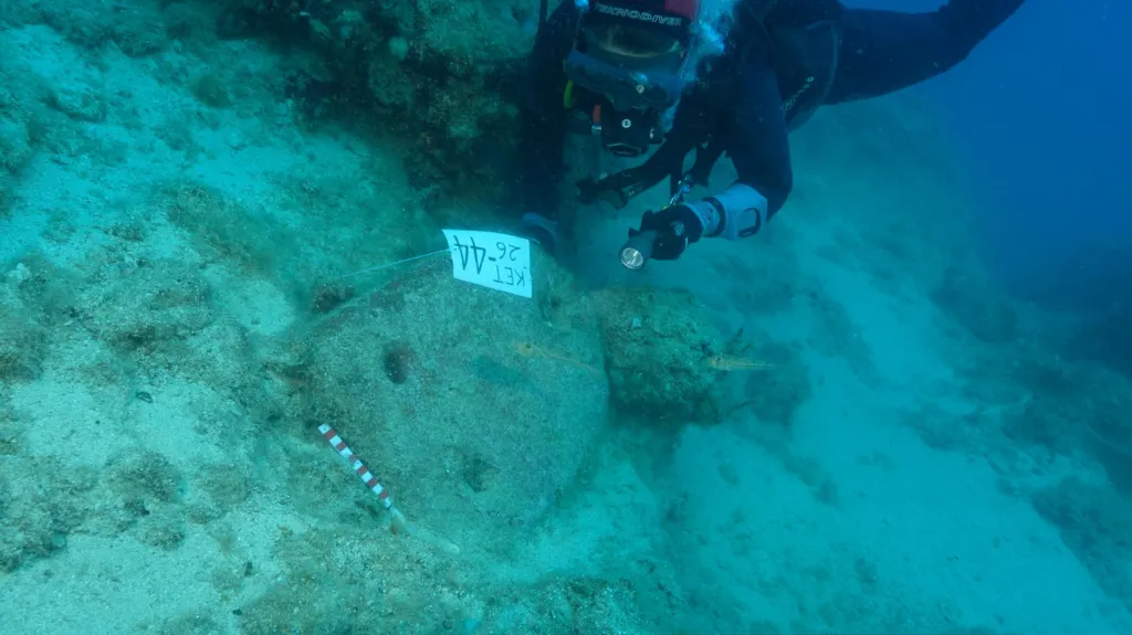 En dykker bruger en lommelygte til at undersøge et stenfund under vandet, der er mærket med en etiket med påskriften KET 44 26.