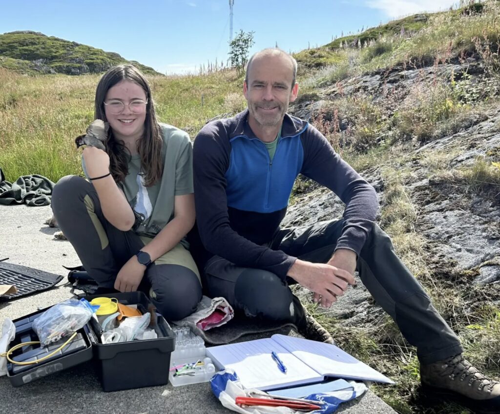 En pige, der holder en lille fugl, og en mand, der sidder på en stenet undergrund udendørs ved siden af feltforskningsudstyr og en notesbog.