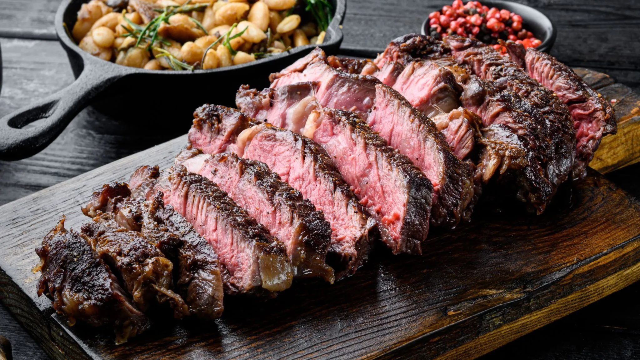 En skåret bøf, medium-rare, på et træskærebræt med en side ret bestående af bønner og peberkorn.