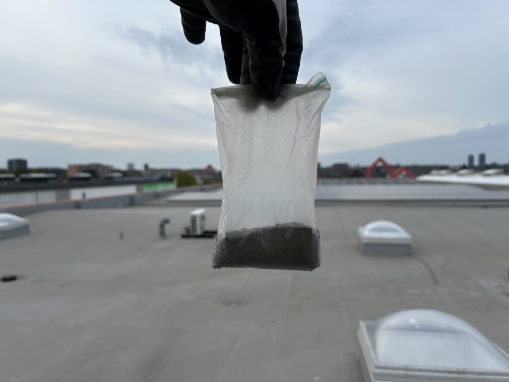En hånd iført en handske holder en lille gennemsigtig plastikpose, der indeholder mørkt pulver, mens et tag og byens skyline ses sløret i baggrunden under en overskyet himmel.