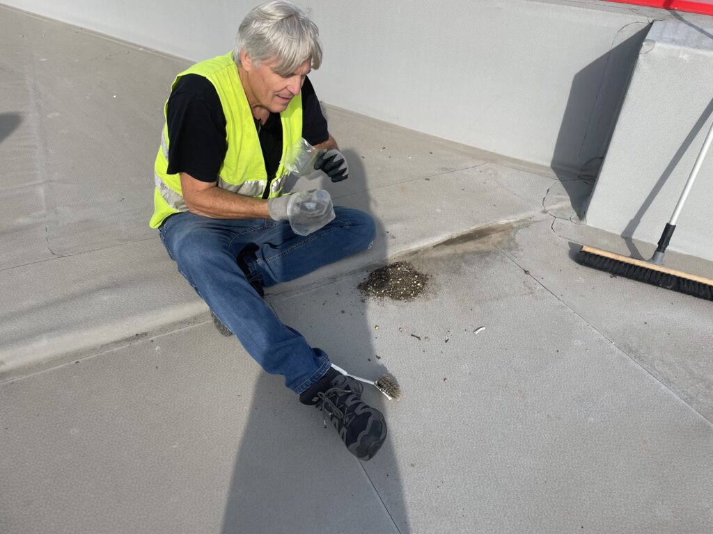En person iført en gul sikkerhedsvest sidder på et tag med en lille beholder i hånden og handsker på, ved siden af en bunke murbrokker og en lille fugl, med en kost og en mur i baggrunden.