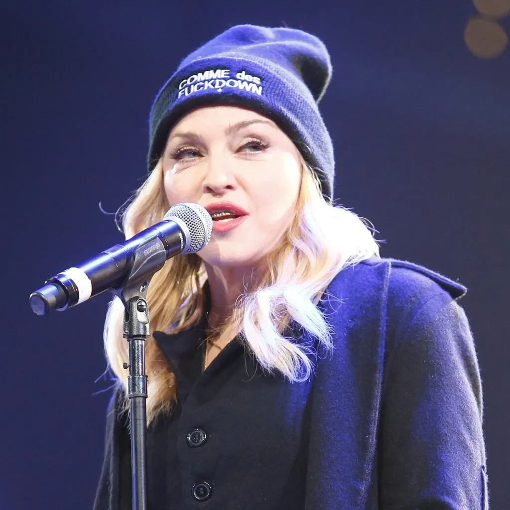 Madonna synger i en mikrofon, mens hun har en sort hue på med påskriften COMME des FUCKDOWN.
