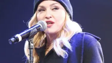 Madonna synger i en mikrofon, mens hun har en sort hue på med påskriften COMME des FUCKDOWN.
