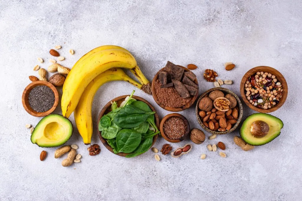 Et flat lay-billede af sunde fødevarer, herunder bananer, halve avocadoer, mørk chokolade, spinat, forskellige nødder, frø og bælgfrugter i træskåle.
