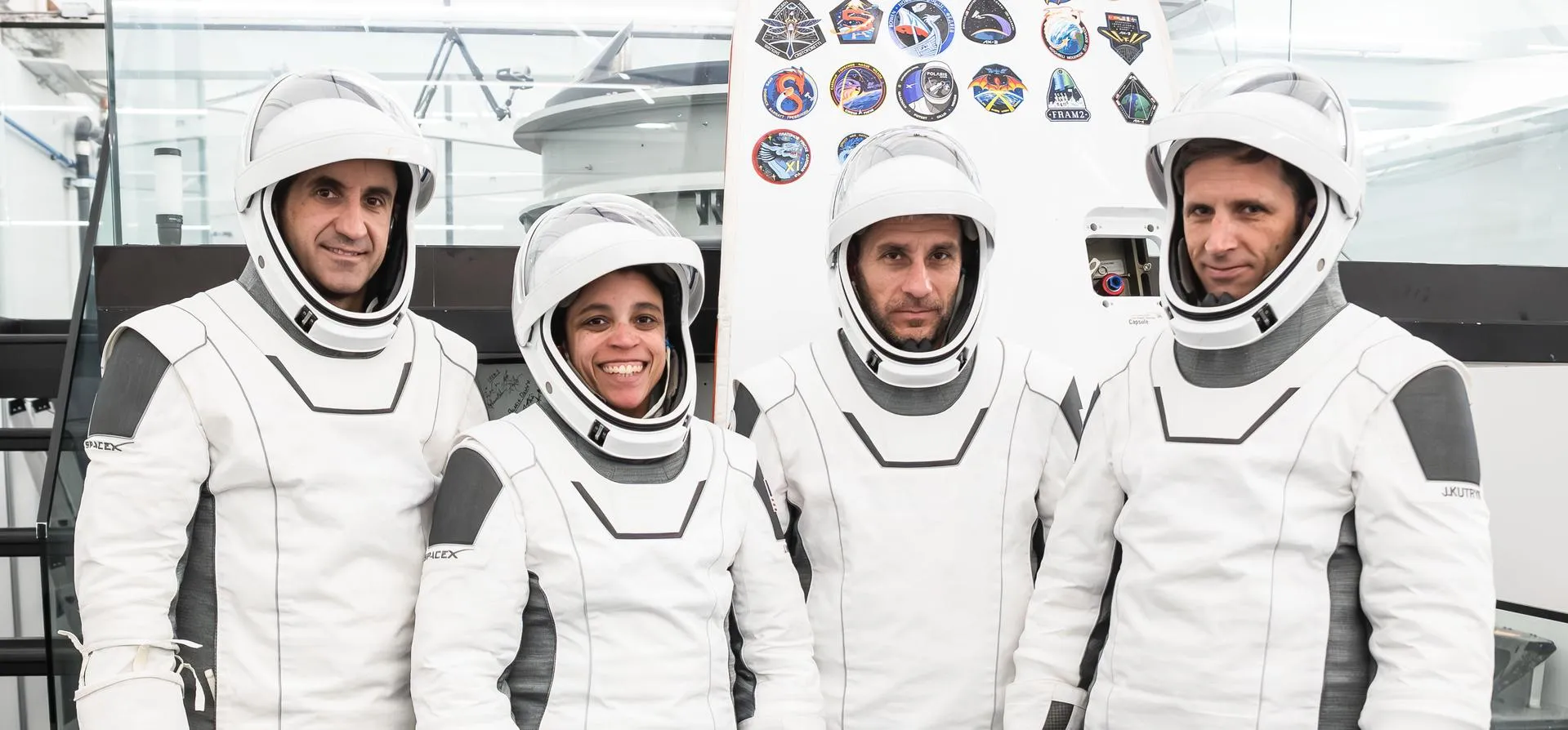 Fire astronauter i hvide SpaceX-dragter står sammen indendørs, smiler og poserer til et foto. Mission-mærker er ophængt på væggen bag dem.