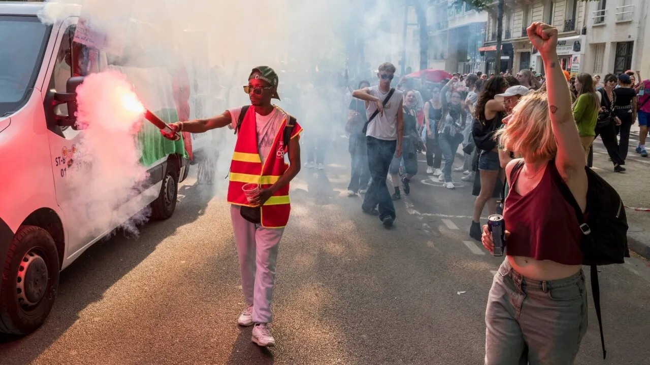 En person iført en refleksvest holder en rød nødraket, mens vedkommende går i en gadedemonstration ved siden af en hvid varevogn og en menneskemængde.